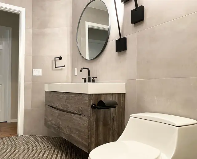 Powder room with a floating wood vanity, round black mirror, black sconce and large-format gray tile